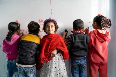 Four children write on a whiteboard with one girl facing the other way smiles at the camera