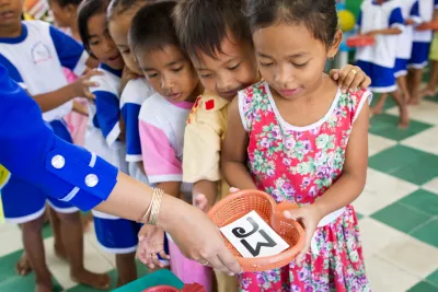 A group of young children stand in a line with the girl at the front of the line holding a piece of paper with Khmer script on it in a basket