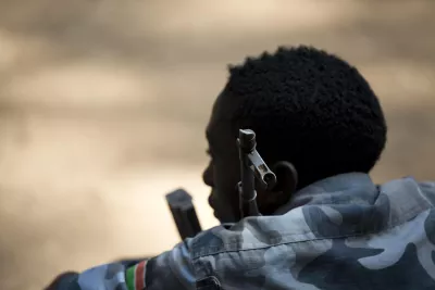 A child soldier in military fatigues sitting down with a gun resting on his shoulder